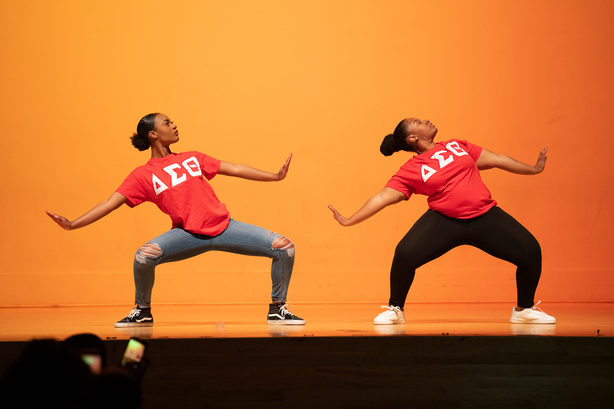 Two women on stage in red shirts with the Delta Sigma Theta logo in white, leaning to the left as they do a step performance in front of a yellow backdrop