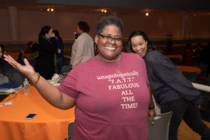 A woman with natural hair smiles at the camera with her right arm stretched out to the side, palm up. Another woman leans into the photo from the right side with a smile, as if photobombing